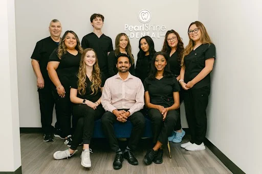 Group photo of eleven dental team members standing and sitting in front of a wall with the 'Pearl Shine Dental' logo. Most are wearing black scrubs, and one person in the center wears a light-colored dress shirt and dark pants.