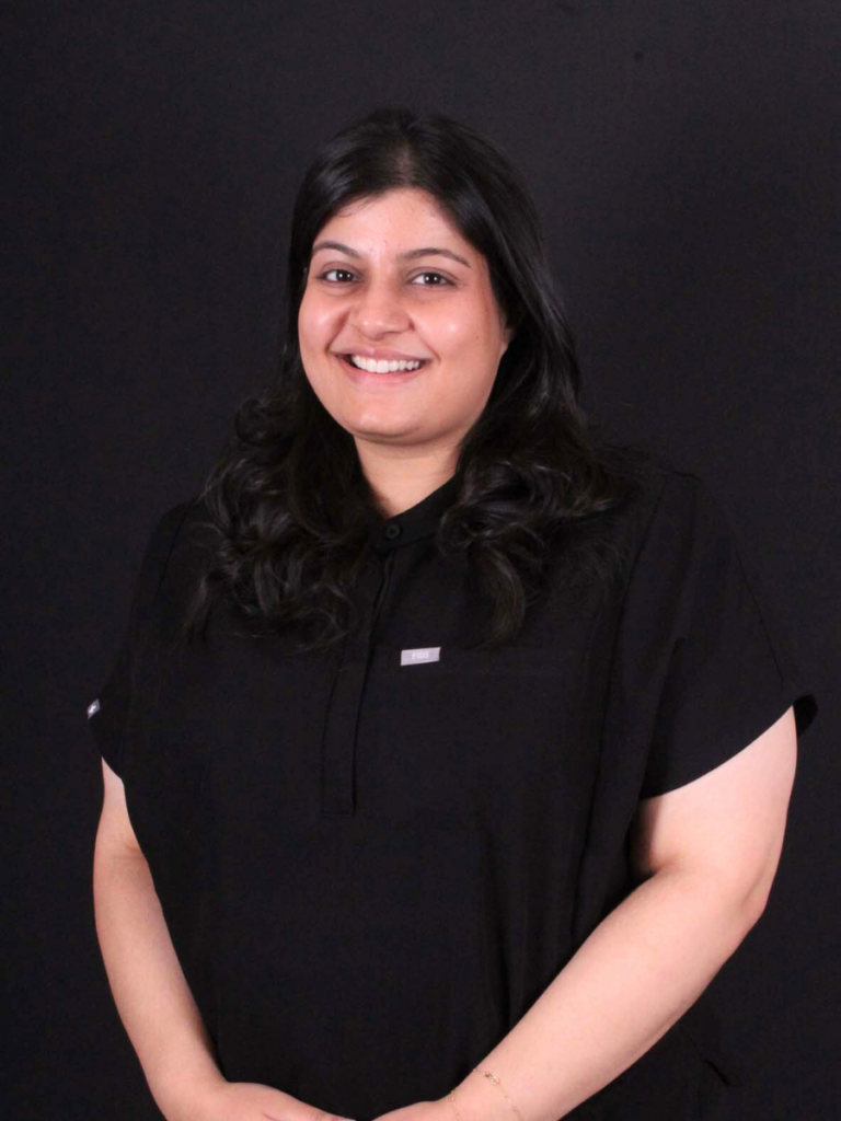 The image shows a person standing against a black background. She was dressed in a black shirt with a small rectangular tag on the chest pocket. Their arms are visible, positioned with hands resting in front.